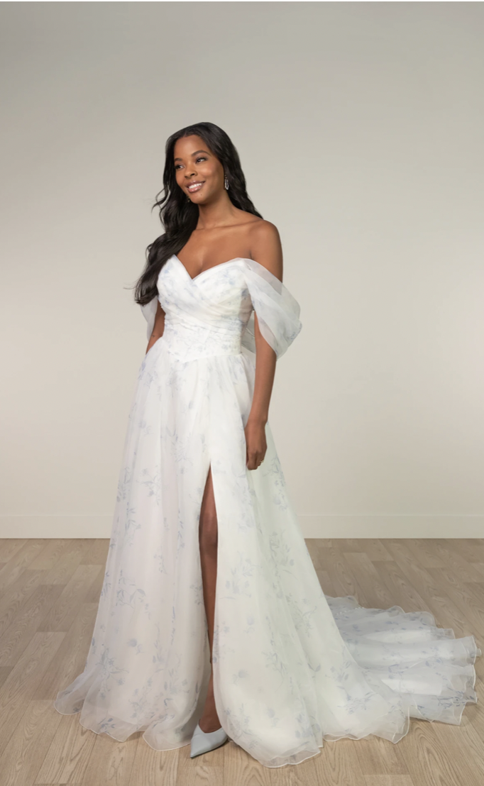 A woman in an elegant, flowing white off-shoulder gown with a floral pattern and train stands smiling. The dress has a thigh-high slit.