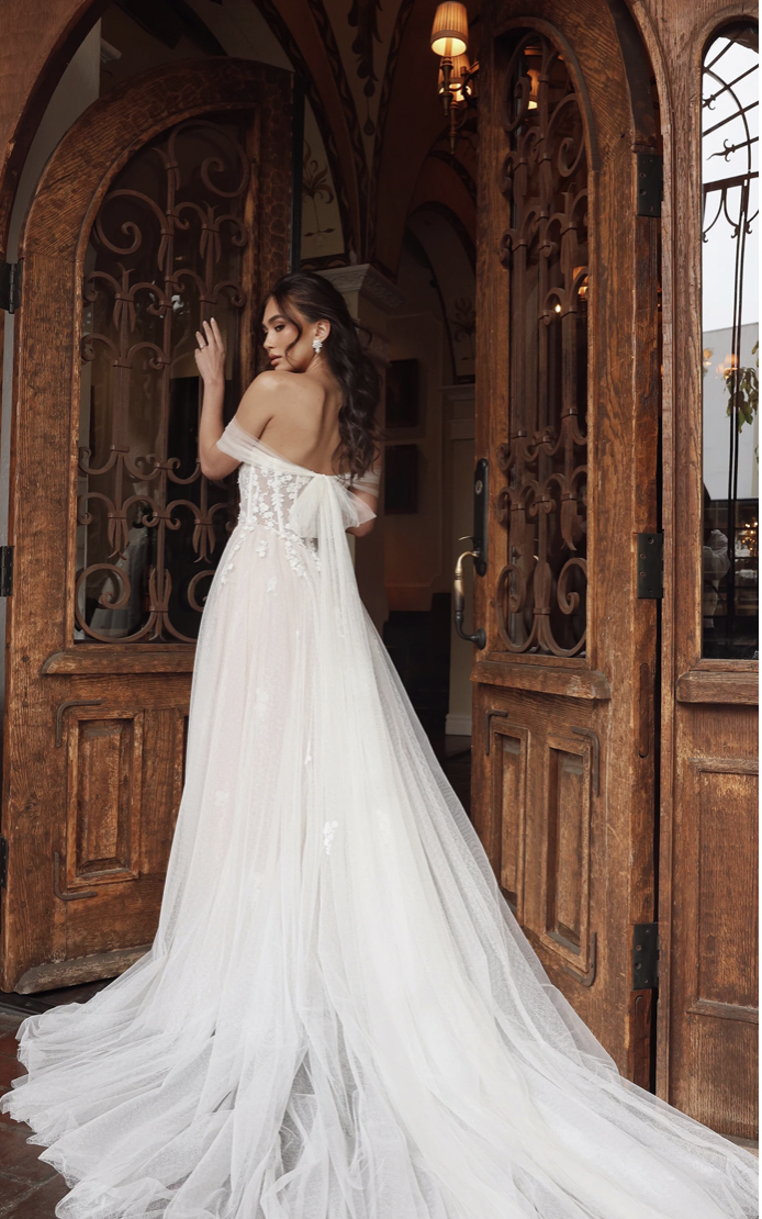 Model is wearing the Essense of Australia Pilar gown, a romantic A-line dress with sequin floral lace, floaty tulle, and a plunging off-the-shoulder neckline.