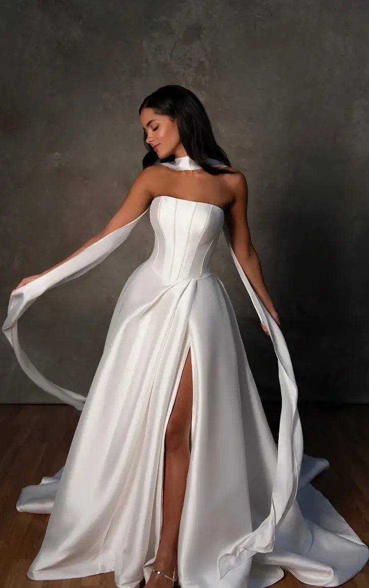 Woman in an elegant, flowing white gown with a slit and long, flowing accents stands on a wooden floor against a textured gray backdrop, exuding grace.