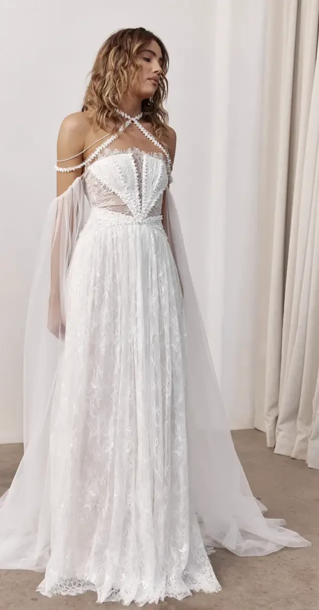 A woman wearing an elegant white dress with intricate detailing and sheer cape-like sleeves.