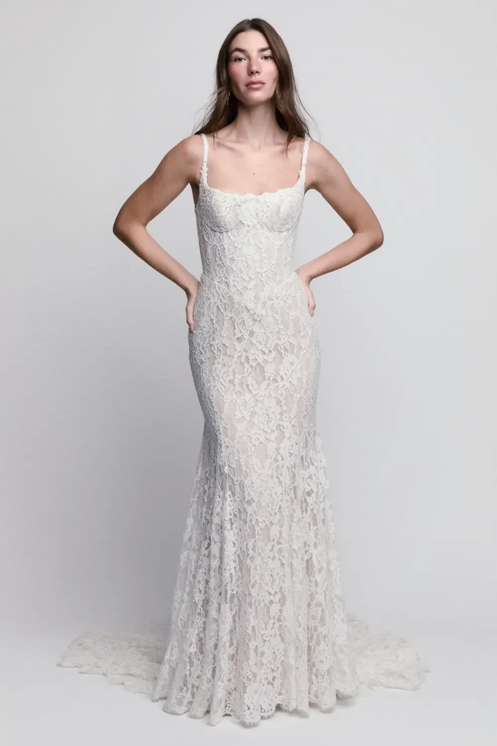 A model wearing a fitted, lace wedding gown with thin straps, standing with hands on hips against a plain background.