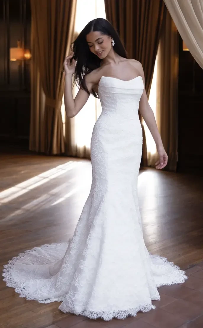 A woman in a strapless white wedding dress stands in a beautifully lit room with wooden floors and elegant drapes.