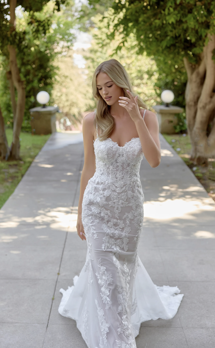 A woman in a lace wedding dress walks down a sunlit path, flanked by green trees, exuding elegance and tranquility with a serene expression.
