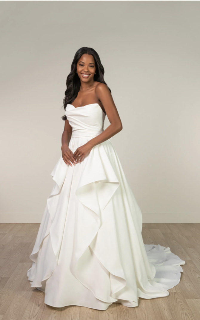 A woman stands in a strapless white wedding gown with a layered skirt. Her long, dark hair is styled in waves. She smiles warmly in a minimalist setting.