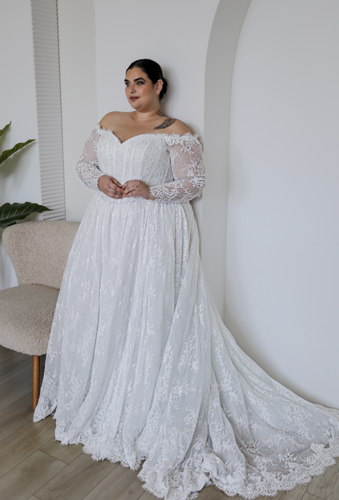 A plus-size woman wearing a long-sleeve white wedding dress with lace detailing, standing next to a beige chair and in front of a white wall.