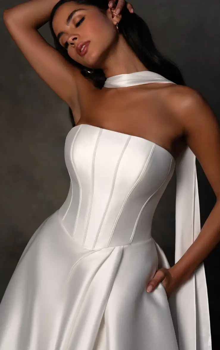 A woman wearing a strapless, white satin dress with a fitted bodice and flowing skirt, posing elegantly with one hand on her hip and the other raised to her hair.