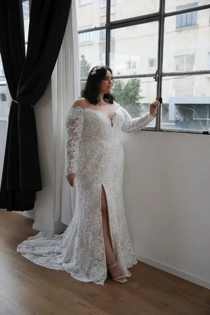 A woman in a white lace wedding dress with off-the-shoulder sleeves, standing by a large window. The dress features a thigh-high slit and she has long, dark hair.